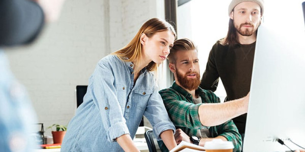 Three young professionals—a woman in a denim shirt, a bearded man in a plaid shirt, and a man in a beanie—lean in toward a large computer monitor in a bright office to analyze and discuss a digital project on the screen.