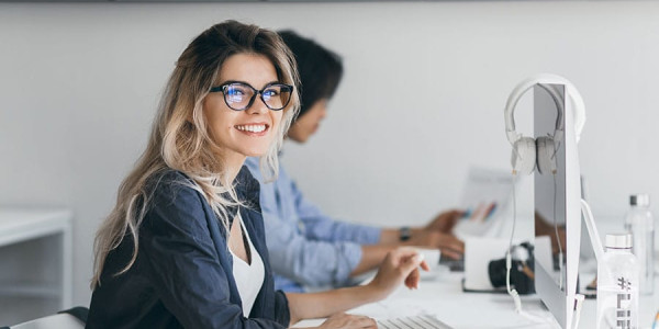 A smiling woman with blonde hair and black-rimmed glasses sits at a white desk in a minimalist office, with a computer monitor, keyboard, and a cup of coffee in front of her as she prepares to write digital content.