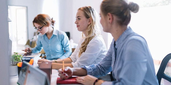 Three professional women sit at a long white desk in a bright office; one woman in a blue shirt uses a tablet, while another in a striped shirt takes notes in a red planner as they collaborate on website budget calculations.
